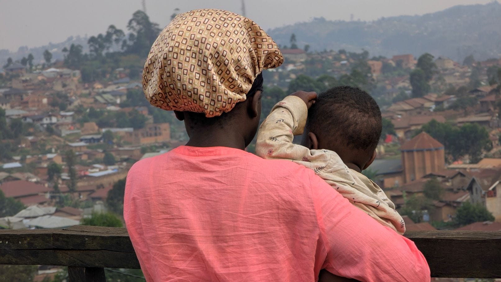 A Congoloese woman wearing a pink dress and patterned head scarf holds a baby. Both are facing away, looking into a ruined landscape