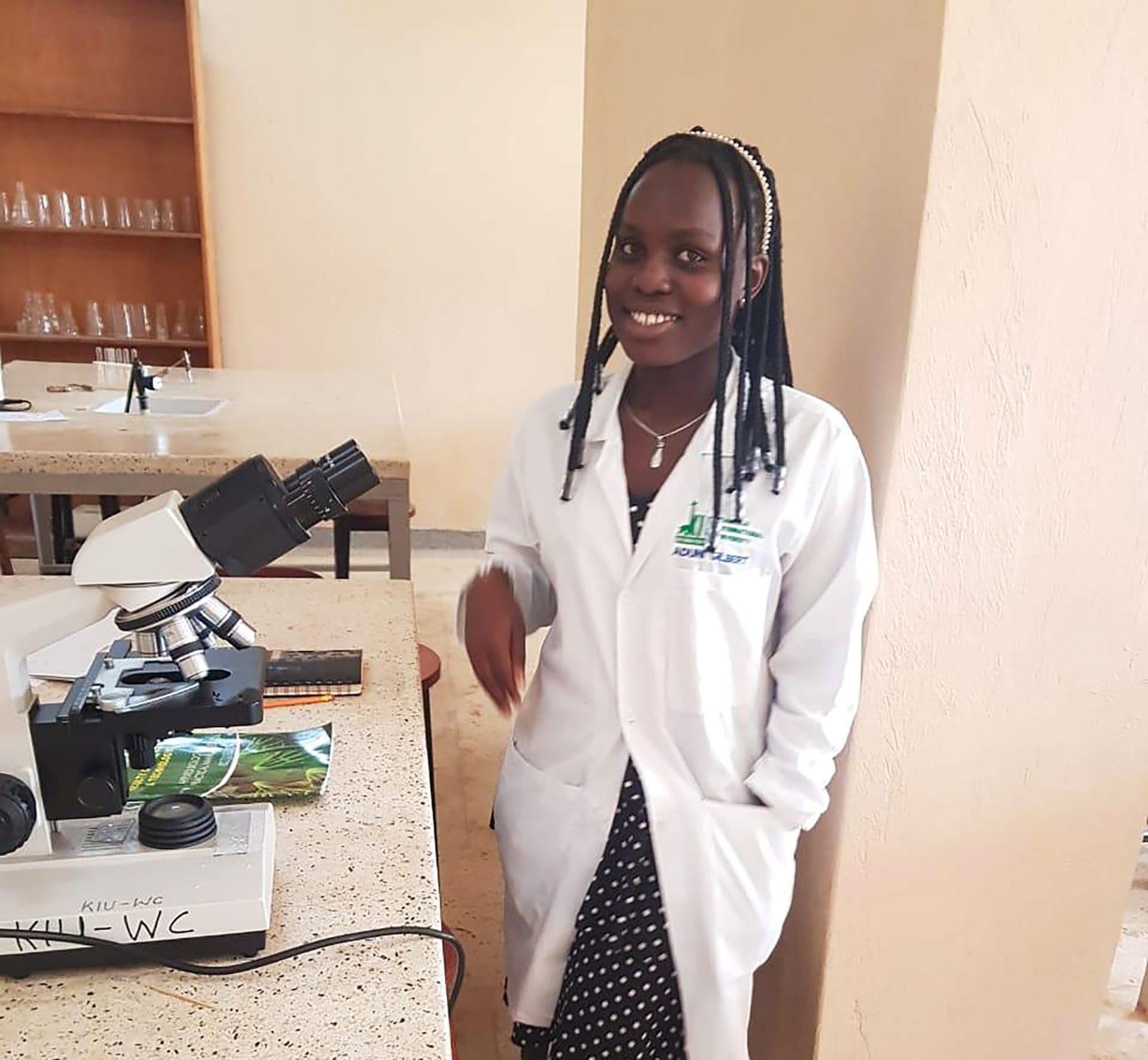 Medical student Hellen Asio wears a white lab coat. Young, black woman with long braided hair. Standing next to lab equipment.